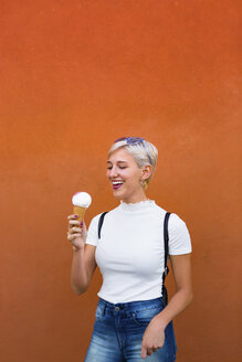 Lachende junge Frau mit Eiswaffel vor orangefarbenem Hintergrund - MGIF00259