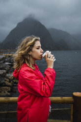 Norwegen, Lofoten, junge Frau an der Küste, die aus einem Kaffee zum Mitnehmen trinkt - KKAF01880