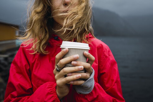 Norwegen, Lofoten, Nahaufnahme einer jungen Frau an der Küste mit Kaffee zum Mitnehmen - KKAF01881