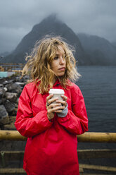 Norwegen, Lofoten, junge Frau an der Küste mit Kaffee zum Mitnehmen - KKAF01882