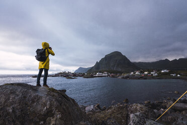 Norwegen, Lofoten, Rückansicht eines Mannes, der auf einem Felsen an der Küste steht und ein Foto macht - KKAF01885