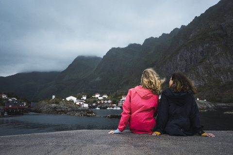 Norwegen, Lofoten, Rückansicht von zwei jungen Frauen, die auf einem Steg an der Küste sitzen, lizenzfreies Stockfoto