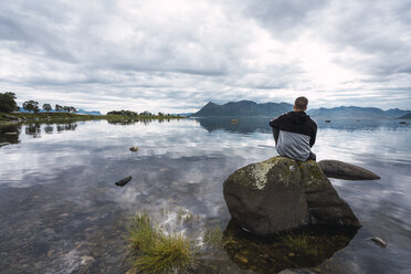 Norwegen, Insel Senja, Rückansicht eines Mannes, der auf einem Felsen an der Küste sitzt - KKAF01901