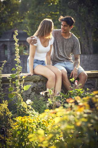 Romantisches junges Paar, das im Park auf einer Mauer sitzt und die Arme umeinander legt, lizenzfreies Stockfoto