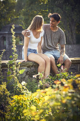 Romantisches junges Paar, das im Park auf einer Mauer sitzt und die Arme umeinander legt - SRYF00787