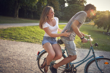 Young couple riding bicycle in park, woman sitting on rack - SRYF00845