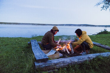 Sweden, Lapland, Two friends preparing a bonfire at the lakeshore - KKAF01924