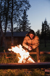 Junger Mann sitzt an einem Lagerfeuer und beobachtet die Flammen - KKAF01929
