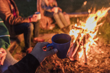 Hand einer Person, die eine Teetasse hält, Gruppe von Menschen, die an einem Lagerfeuer sitzen - KKAF01950