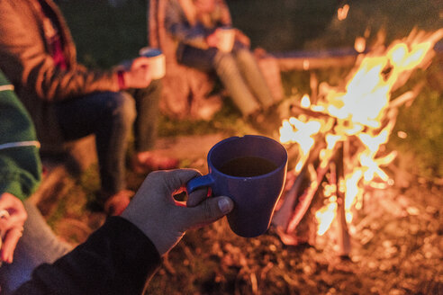 Hand of person holding tea cup, group of people sitting at a camp fire - KKAF01950