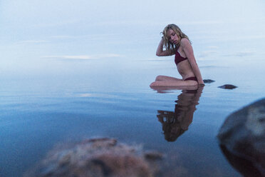 Junge Frau im Bikini, sitzend auf einem Stein in einem See - KKAF01961