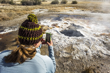 BORAX LAKE, HARNEY COUNTY, OR, USA: Eine junge Frau mit Wollmütze fotografiert mit ihrem Smartphone eine dampfende, kochende heiße Quelle, die mit weißen Kalkablagerungen verkrustet ist. - AURF04906