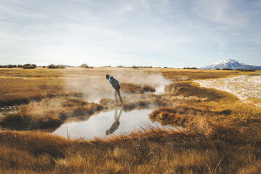 BORAX LAKE, HARNEY COUNTY, OR, USA: Eine junge Frau in einem blauen Mantel beugt sich vor, um in eine dampfende heiße Quelle in einer weiten, trockenen Sumpflandschaft zu blicken. - AURF04907