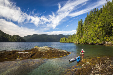 HAIDA GWAII, BRITISH COLUMBIA, KANADA: Ein Mann zieht zwei Kajaks durch seichtes, türkisfarbenes Wasser entlang einer felsigen, bewaldeten Küste. - AURF04963