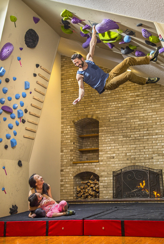 Mutter und Tochter betrachten den Mann, der an der Kletterwand im Indoor-Fitnessstudio im Haus klettert, lizenzfreies Stockfoto