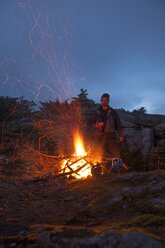 Porträt eines Bergsteigers, der sich neben einem alpinen Lagerfeuer warm hält - AURF05408