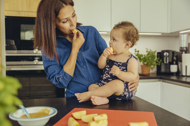 Mutter und kleine Tochter essen gemeinsam Apfelstücke in der Küche - MFF04648