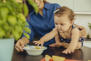 Mother with baby daughter eating fruit pulp in kitchen - MFF04649