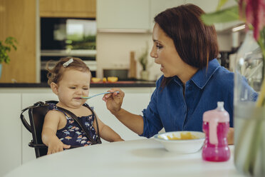 Mother helping disgusted baby daughter eating fruit pulp in kitchen - MFF04653