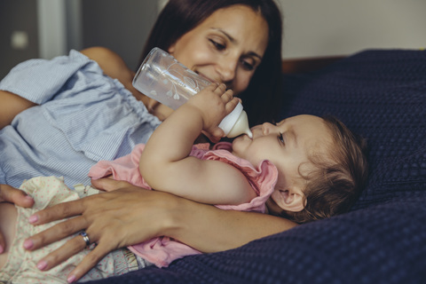 Mother watching her baby girl drinking milk from a bottle on bed stock photo