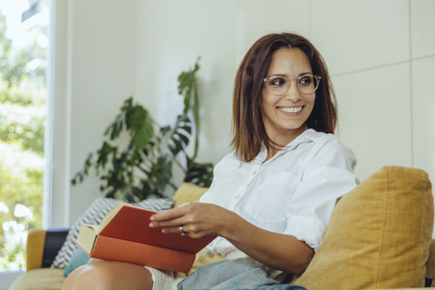 Smiling woman looking up from reading book on couch stock photo