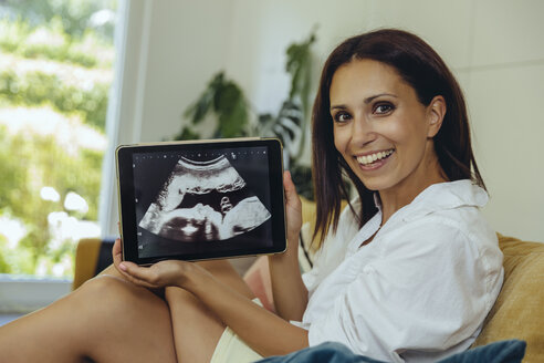 Porträt einer lächelnden Frau, die ein Ultraschallbild ihres ungeborenen Kindes auf einer Couch zeigt - MFF04705