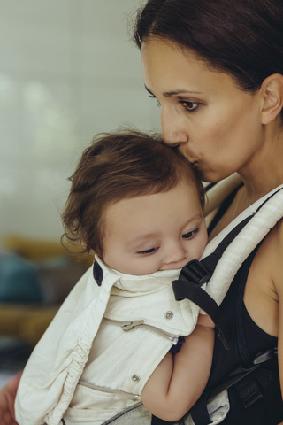 Mutter küsst und trägt ihr kleines Mädchen im Tragetuch nach Hause, lizenzfreies Stockfoto