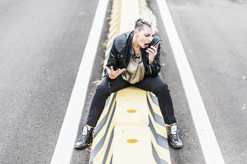 Aggressive Punkerin sitzt am Straßenrand und schreit in ihr Handy, lizenzfreies Stockfoto