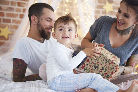 Glücklicher Junge öffnet Weihnachtsgeschenk mit seinen Eltern im Bett, lizenzfreies Stockfoto