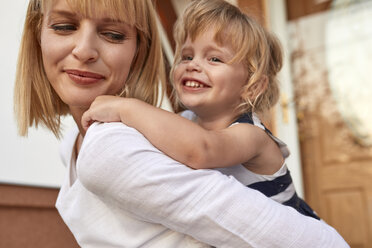 Smiling mother carrying her daughter piggyback in front of their house - ZEDF01573