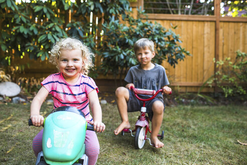 SEATTLE, WA, USA. A smiling blond toddler girl in pink and a blond boy sit on tricycles in a backyard. - AURF05533