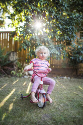 SEATTLE, WA, USA: Ein blondes, pinkfarbenes Kleinkind sitzt auf einem Dreirad in einem Hinterhof, in dem das Sonnenlicht durch die Bäume fällt. - AURF05534