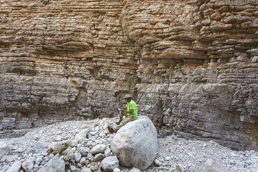 Älterer Mann sitzt auf einem Felsen in einer engen trockenen Schlucht mit steilen, geschichteten Felswänden - AURF05535