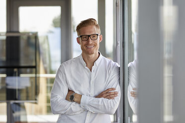 Portrait of smiling businessman in office leaning against window - RBF06708