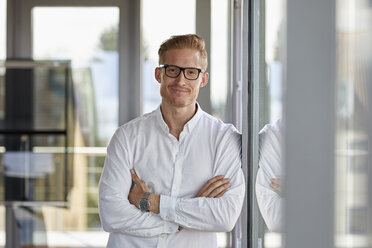 Portrait of smiling businessman in office leaning against window - RBF06713