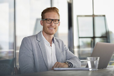 Portrait of smiling businessman with laptop on desk in office - RBF06737