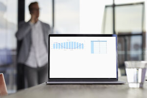 Chart auf Laptop-Bildschirm auf dem Schreibtisch im Büro mit Geschäftsmann im Hintergrund - RBF06743