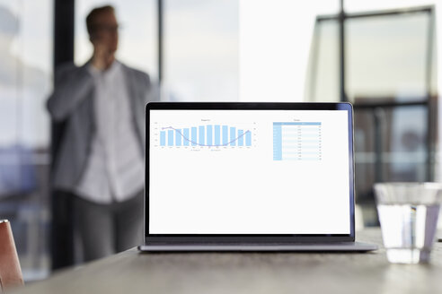 Chart on laptop screen on desk in office with businessman in background - RBF06743
