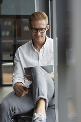 Smiling businessman sitting on windowsill using tablet - RBF06764