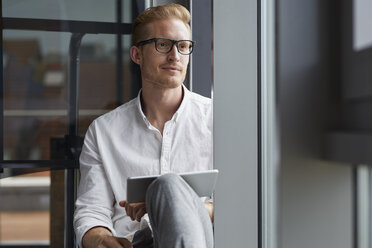 Smiling businessman sitting on windowsill with tablet - RBF06766