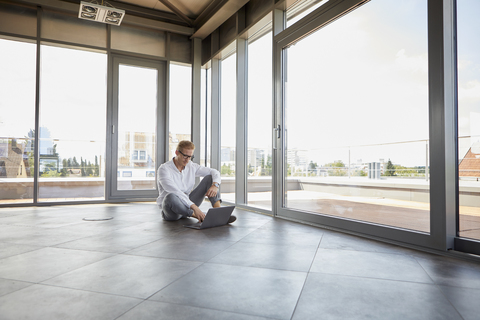 Geschäftsmann sitzt in einem leeren Raum mit Panoramafenster und benutzt einen Laptop, lizenzfreies Stockfoto