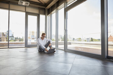 Businessman sitting in empty room with panorama window using laptop - RBF06772