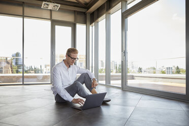 Businessman sitting in empty room with panorama window using laptop - RBF06773