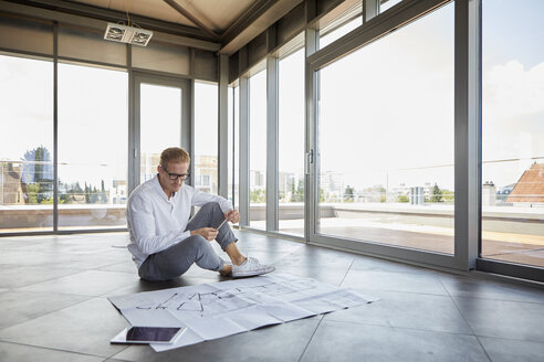 Junger Mann sitzt in leerem Raum mit Panoramafenster und arbeitet an einem Bauplan - RBF06774