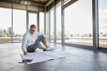 Junger Mann sitzt in leerem Raum mit Panoramafenster und arbeitet an einem Bauplan - RBF06775