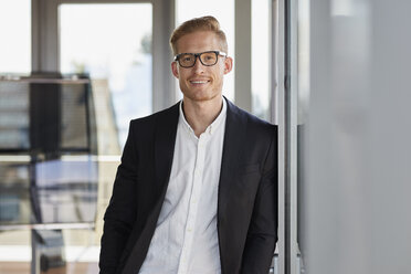 Portrait of smiling businessman in office leaning against window - RBF06777