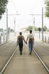 Rückenansicht eines jungen schwulen Paares mit Rucksäcken, das Hand in Hand zwischen den Gleisen auf der Straße läuft - FBAF00048