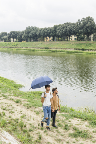 Junges schwules Paar mit Regenschirm spaziert Hand in Hand am Flussufer, lizenzfreies Stockfoto