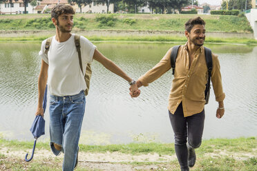 Portrait of young gay couple with backpacks walking hand in hand at riverside - FBAF00050