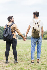 Rückenansicht eines jungen schwulen Paares mit Rucksäcken, das Hand in Hand auf einer Wiese steht - FBAF00060